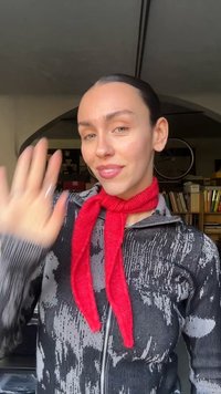 Juoda užsegama striukė su tekstūruotu raštu, derinama su ryškiai raudonu šaliku, surištu ant kaklo. Neutrali fonas su matomomis lentynomis.