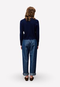 Persoon met schouderlang haar draagt een marineblauwe geribbelde trui, losse opgerolde blauwe spijkerbroek en bruine instapschoenen, staande met de rug naar de kijker.