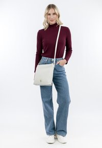 Borsa a tracolla bianca con una texture liscia e dettagli in hardware dorato. Modella che indossa un dolcevita bordeaux a coste e jeans blu a vita alta.