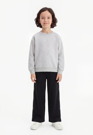 Barn med kort mørkt hår iført en lysegrå sweatshirt, sorte bukser med vide ben og knapper i siden samt hvide sneakers, står på en hvid baggrund.