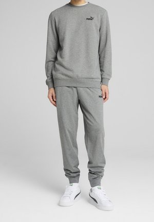 Person trägt grauen Puma-Sweatshirt und passende Jogginghose mit weißen Puma-Sneakers, steht vor schlichtem grauen Hintergrund.