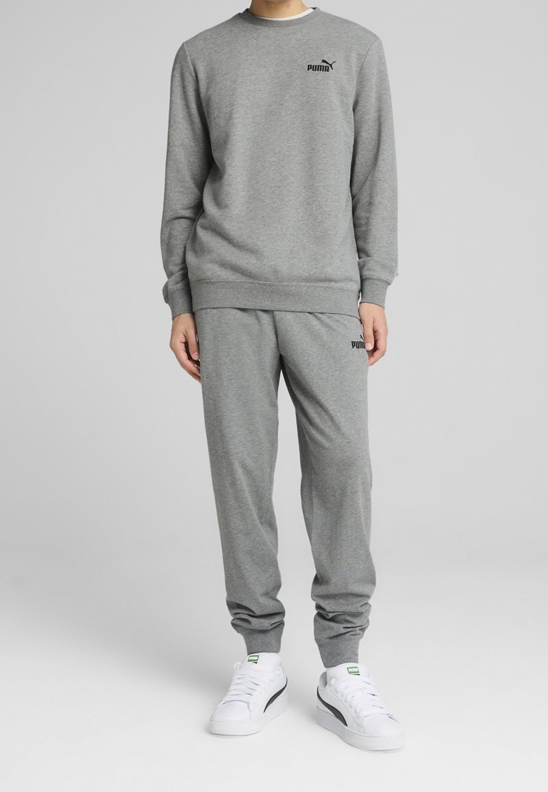 Personne portant un sweat-shirt gris Puma et un pantalon de survêtement assorti avec des baskets blanches Puma, debout devant un fond gris uni.
