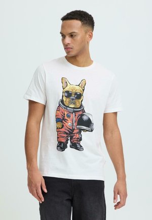 Jeune homme portant un t-shirt blanc avec un bouledogue français en combinaison d'astronaute orange et lunettes de soleil, tenant un casque spatial noir.