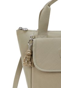 Bolso de nailon beige con solapa frontal, cremallera plateada y un llavero de mono de plástico añadido. Textura suave, interior completamente forrado, correa ajustable.