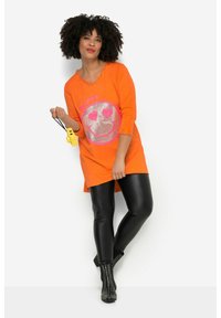 Orange Baumwolltunika mit einem silbernen Glitzer-Smiley-Gesicht und pinkem Text "HAPPY", kombiniert mit schwarzen glänzenden Leggings und mit Nieten besetzten Ankle Boots.
