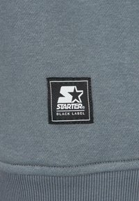Grauer Sweatshirt-Stoff mit einem rechteckigen schwarzen Etikett, auf dem der weiße Text "STARTER" und das Stern-Logo zu sehen sind. Gerippte Bündchendetails am Saum.