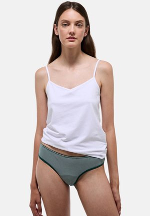 Jeune femme aux longs cheveux bruns porte un caraco blanc et un sous-vêtement rayé vert, debout devant un fond clair uni.