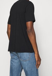 Person iført en ensfarvet sort kortærmet t-shirt og lyseblå denimjeans, vist fra ryggen mod en ensfarvet hvid baggrund.