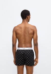 Μαύρα ανδρικά μποξέρ με λευκό λογότυπο CK και μικρό λευκό μοτίβο, με ελαστική μέση. Το μοντέλο είναι γυρισμένο πλάτη, παρουσιάζοντας την πίσω όψη και το σχέδιο των σορτς.