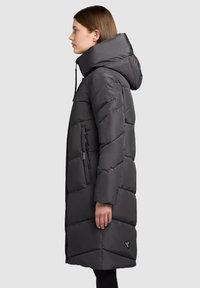 Jachetă matlasată gri cu guler înalt și glugă mare. Prezintă un model matlasat diagonal, buzunare cu fermoar și un accent cu logo la tiv.