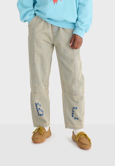 Personne portant un jean beige avec des motifs carrés bleus près des genoux, un sweat-shirt bleu clair, des chaussettes blanches et des mocassins marron avec des lacets jaunes.