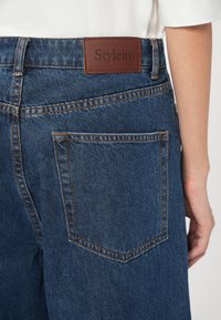 Mørkeblå denimjeans med et brunt lærmerke som leser "Stylein" i midjen. Har dobbel søm og baklommer.