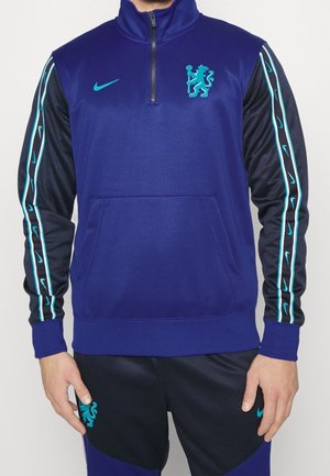 Mann trägt eine blau-schwarze Nike-Sportjacke und Hose mit türkisfarbenem Löwenwappen und Nike Swoosh-Logos.
