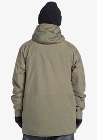 Olivengrøn vandtæt jakke med hætte, der har zip-lukning halvvejs ned, elastiske manchetter og sorte handsker. Glat ydre med mat finish.