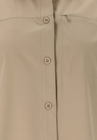 Beige overhemd voorkant met drie zichtbare knopen en een ritssluiting op de borstzak aan de rechterkant. Glad stof met zichtbare stiksels.