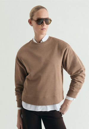 Mujer vistiendo una sudadera marrón de gran tamaño sobre una camisa blanca, pantalones oscuros y grandes gafas de sol marrones, de pie con una mano en la cadera.