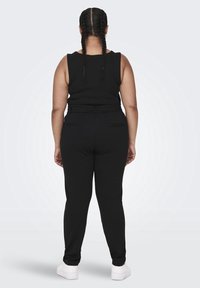 En person med flätat hår står vänd bort, iförd en svart ärmlös topp, svarta byxor och vita sneakers mot en enfärgad bakgrund.