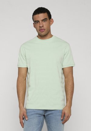 Giovane uomo con indosso una camicia verde chiaro a maniche corte e jeans blu, in piedi davanti a uno sfondo grigio chiaro uniforme.