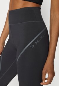 Leggings negros de cintura alta fabricados con un material suave y elástico. Presentan una franja diagonal gris con los números 55, 95, 09, 14 impresos en el lado.
