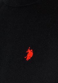Logotipo bordado en rojo de un jugador de polo montado a caballo sobre un fondo de tela ribeteada negra.