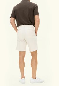 Pantaloni corti bianchi in cotone con vestibilità sartoriale, tasche posteriori e una texture liscia, abbinati a sneaker bianche e a un polo corto a maniche corte scuro.