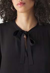 Blouse noire avec une ouverture en forme de clé, présentant un détail à nouer au niveau du col. Texture lisse et accents cousus subtils autour du col.