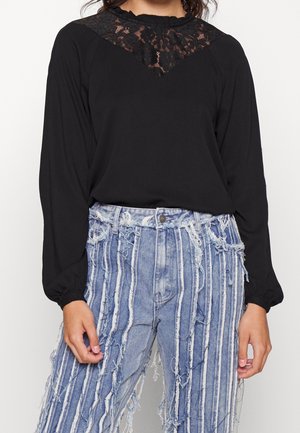 Vrouw draagt een zwart langärmelig shirt met kanten inzetstuk en een high-waist blauwe spijkerbroek met verticale witte strepen en rafelige randen.