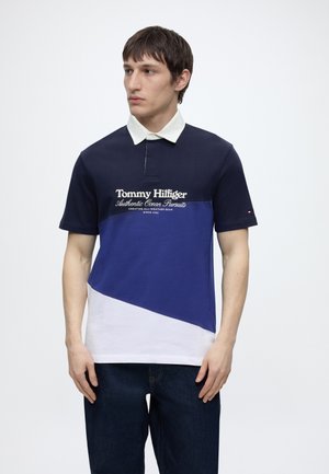 Ung mand iført en farveblokkeret sort, blå og hvid Tommy Hilfiger polo med krave, stående mod en ensfarvet hvid baggrund.