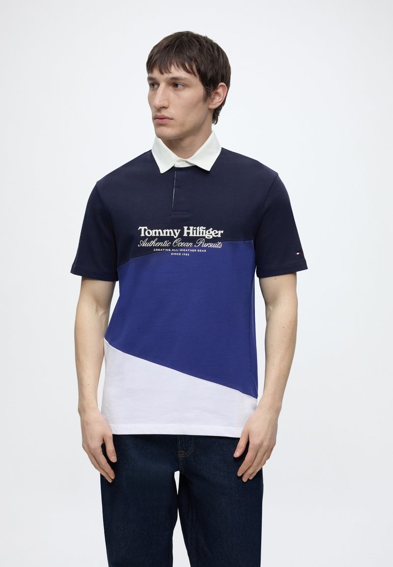 Νεαρός άνδρας που φοράει ένα πόλο Tommy Hilfiger με χρωματικά μπλοκ σε μαύρο, μπλε και λευκό με γιακά, στέκεται μπροστά σε απλό λευκό φόντο.
