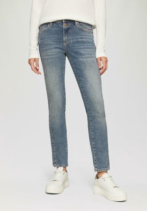 Vrouw draagt lichtblauwe slim-fit jeans, wit shirt met lange mouwen en witte platformsneakers, staand tegen een effen achtergrond.