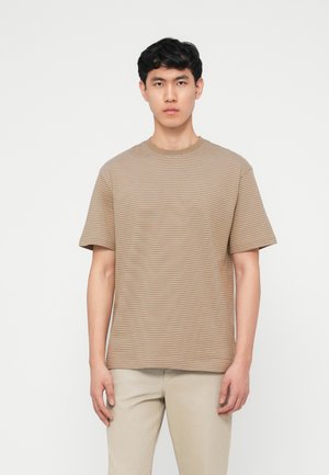 Ung mand stående, iført en beige og hvidstribet T-shirt med korte ærmer og lysebeige bukser mod en ensfarvet hvid baggrund.
