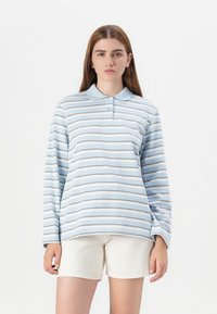 Model with long brown hair wearing a blue and white striped long-sleeve polo shirt and beige shorts, standing against a plain white background.