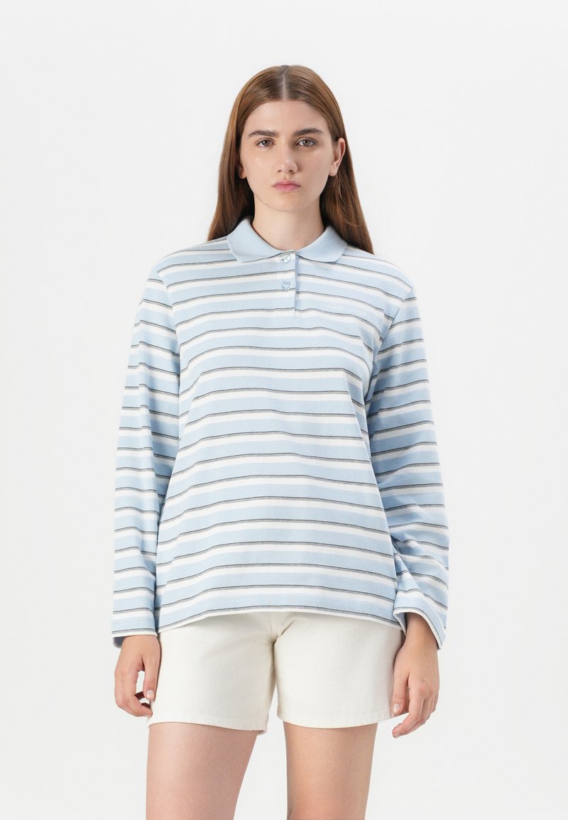 Model with long brown hair wearing a blue and white striped long-sleeve polo shirt and beige shorts, standing against a plain white background.