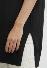 Svart stickad topp med sidsprund, i kombination med strukturerade krämfärgade kordbyxor. Vist är en hand som vilar på plaggets nederkant.