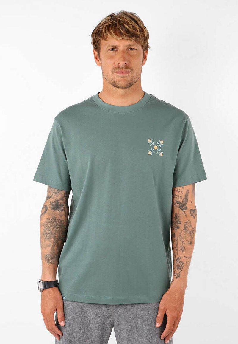 T-shirt en coton vert avec un col rond et des manches courtes, présentant un motif décoratif jaune et bleu sur le côté gauche de la poitrine.