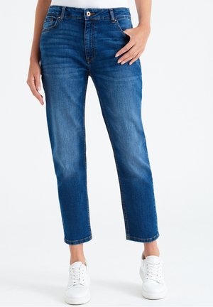 Personne portant un jean denim bleu taille moyenne avec une coupe droite et des baskets blanches, debout avec une main près de la poche.