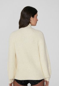 Suéter de punto color crema con un cuello alto, textura acanalada y hombros caídos, que presenta un ajuste relajado y aberturas en los laterales del dobladillo.
