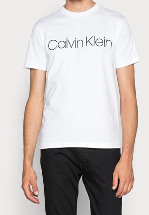 Mężczyzna w białej koszulce z krótkim rękawem z napisem „Calvin Klein” na piersi oraz w czarnych spodniach.