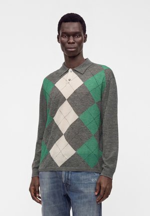 Jeune homme portant un pull à losanges gris, vert et blanc avec un col et un jean bleu, debout devant un fond clair uni.