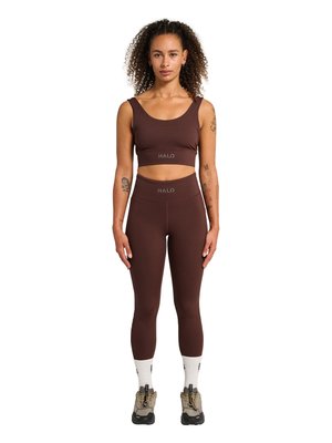 Frau mit lockigem Haar, die ein braunes bauchfreies Oberteil und passende Leggings mit dem Schriftzug „HALO“, weiße Socken und beige Turnschuhe trägt, steht frontseitig.