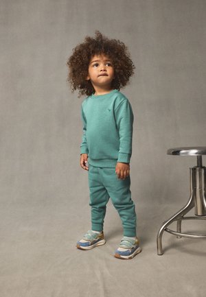 Turkis bomulds-sweatshirt og joggingbukser med ribstrikkede manchetter. Sneakers har blå, grå og gule accenter. Robust metalhynde i nærheden.