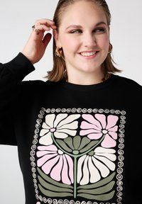 Personne souriante portant des boucles d'oreilles en or en forme de créoles et un haut noir à grand imprimé floral stylisé en tons de rose, blanc et vert.