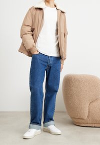 Ljust brunt puffer-jacka med fårskinnskrag, vit t-shirt, blå raka jeans och vita sneakers. Avslappnad passform.