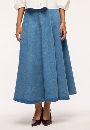 Donna che indossa una gonna midi a trapezio in denim blu medio, una camicia bianca e décolleté scuri con punta affilata, in piedi davanti a uno sfondo uniforme.
