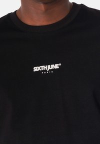 Camiseta negra de cuello redondo hecha de algodón, con el logo bordado en blanco "SIXTHJUNE PARIS" en la parte frontal. Diseño simple y ajustado.