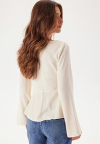 Långärmad vit blus med figursydd överdel och peplum-rygg. Mjuk tyg med subtil textur. Kombineras med högmidjade blå jeans.
