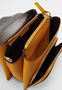 Żółta torba ze skóry z fakturą i brązowymi akcentami, posiadająca trzy zamykane na suwak przegródki oraz zatrzask na górze.