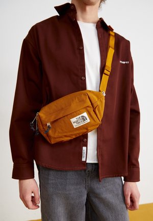 Personne portant une chemise marron à boutons par-dessus un t-shirt blanc avec un sac bandoulière North Face jaune moutarde sur la poitrine.