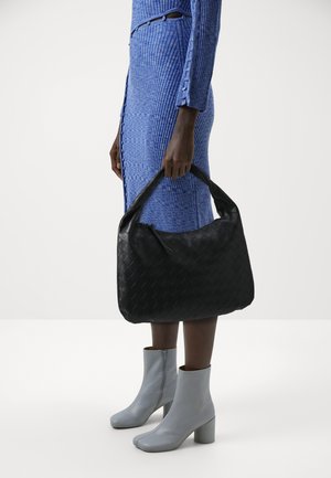 Femme portant une robe bleue côtelée et des bottines grises, tenant un grand sac à main en cuir tressé noir par la poignée.