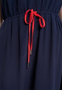 Robe bleu marine avec taille froncée attachée par un nœud coulissant rouge avec embouts métalliques aux extrémités.